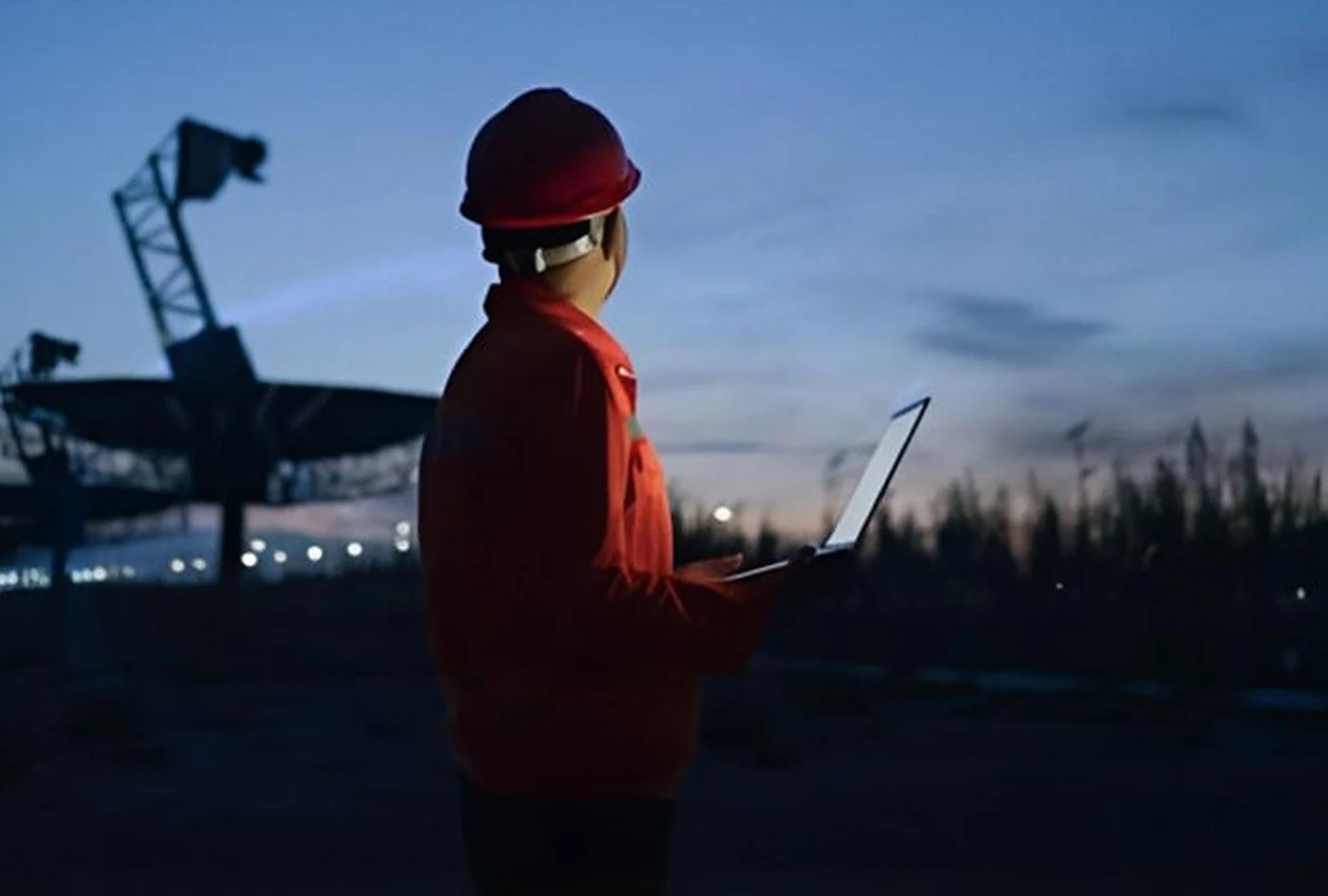 Engineer in a hard hat checking a laptop at an industrial site at dusk